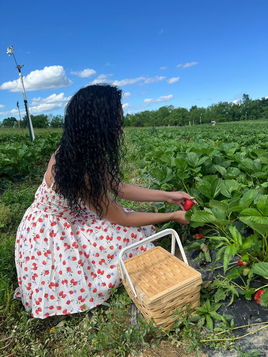 Woman with long, wet curly hair wearing a strawberry-print dress and picking strawberries in a field, showing defined hydrated curls.